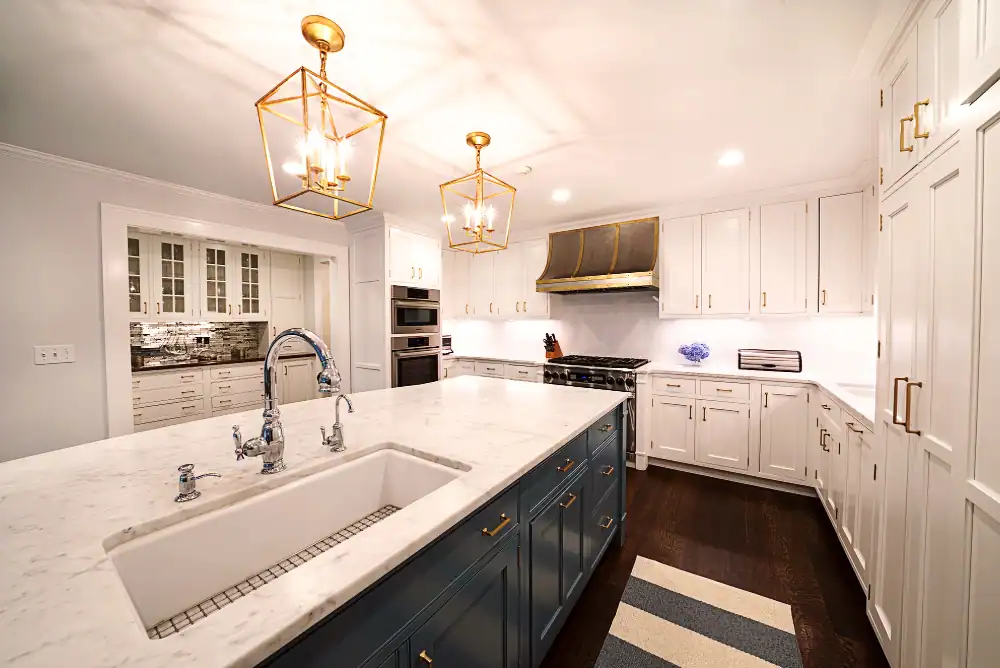 Spacious modern kitchen with white cabinets, a large marble island with a sink, gold pendant lights, dark wood floors, stainless steel appliances, and a striped rug.