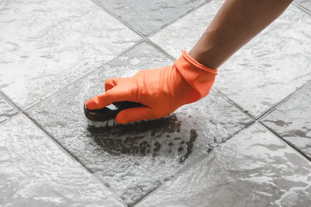 A person wearing an orange glove scrubs the grout between gray floor tiles with a small cleaning brush on a wet surface.